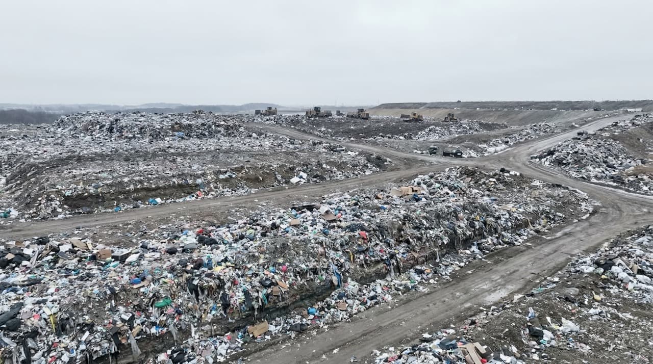 Aerial view of a large commercial landfill site under overcast skies, showing compacted waste layers and distant heavy machinery — representing the scale of the global waste disposal crisis, with true costs estimated at $361 billion annually by the UNEP Global Waste Management Outlook 2024.