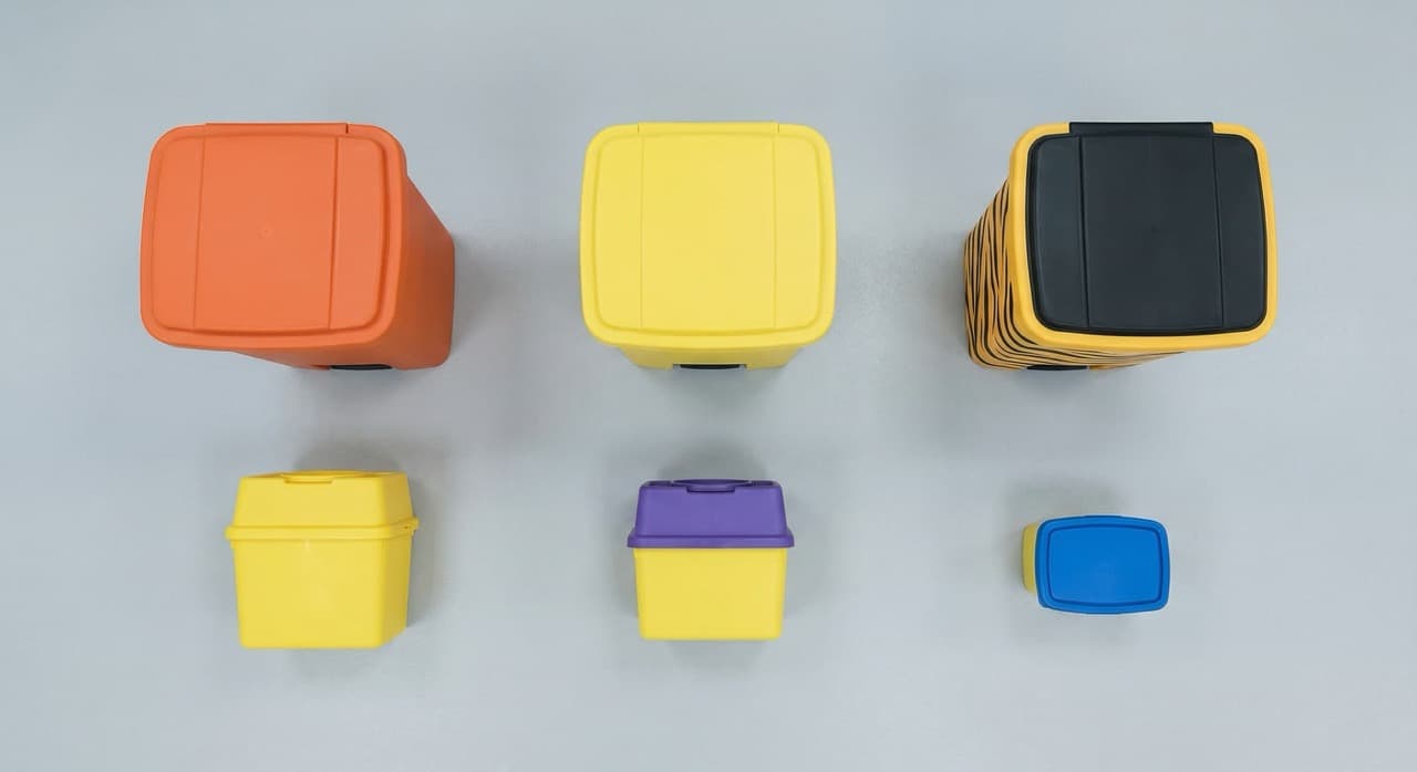 Overhead flat-lay of six NHS clinical waste containers arranged systematically: orange bag holder (Category A infectious waste), yellow bag holder (medicinally contaminated), tiger-stripe bag holder (Category G offensive waste), yellow-lidded sharps container, purple-lidded cytotoxic container, and blue-lidded pharmaceutical container — representing the full HTM 07-01 classification system under UK healthcare waste law.