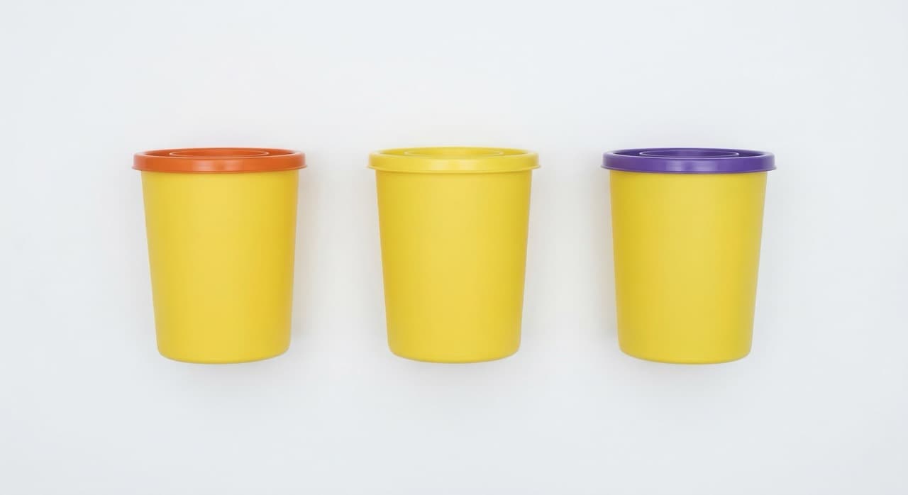 Overhead flat-lay of three NHS clinical sharps containers: orange-lidded (alternative treatment eligible, ~£419/tonne), yellow-lidded (high-temperature incineration mandatory, ~£617/tonne), and purple-lidded (cytotoxic/cytostatic — incineration only). Defaulting to yellow when orange is correct costs £198/tonne more on every sharps disposal cycle.
