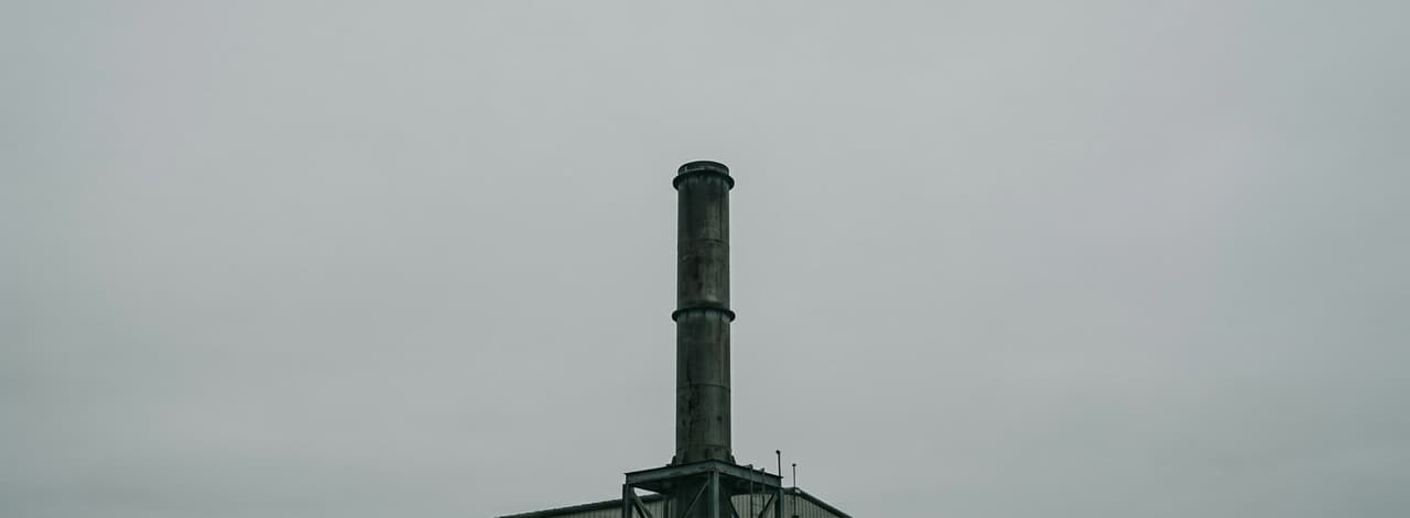 Industrial incineration chimney stack against a flat overcast sky — the combustion infrastructure that generates dioxins, NOₓ, and toxic ash, representing the emission pathway that subcritical water hydrolysis eliminates by operating in a sealed oxygen-free liquid environment.