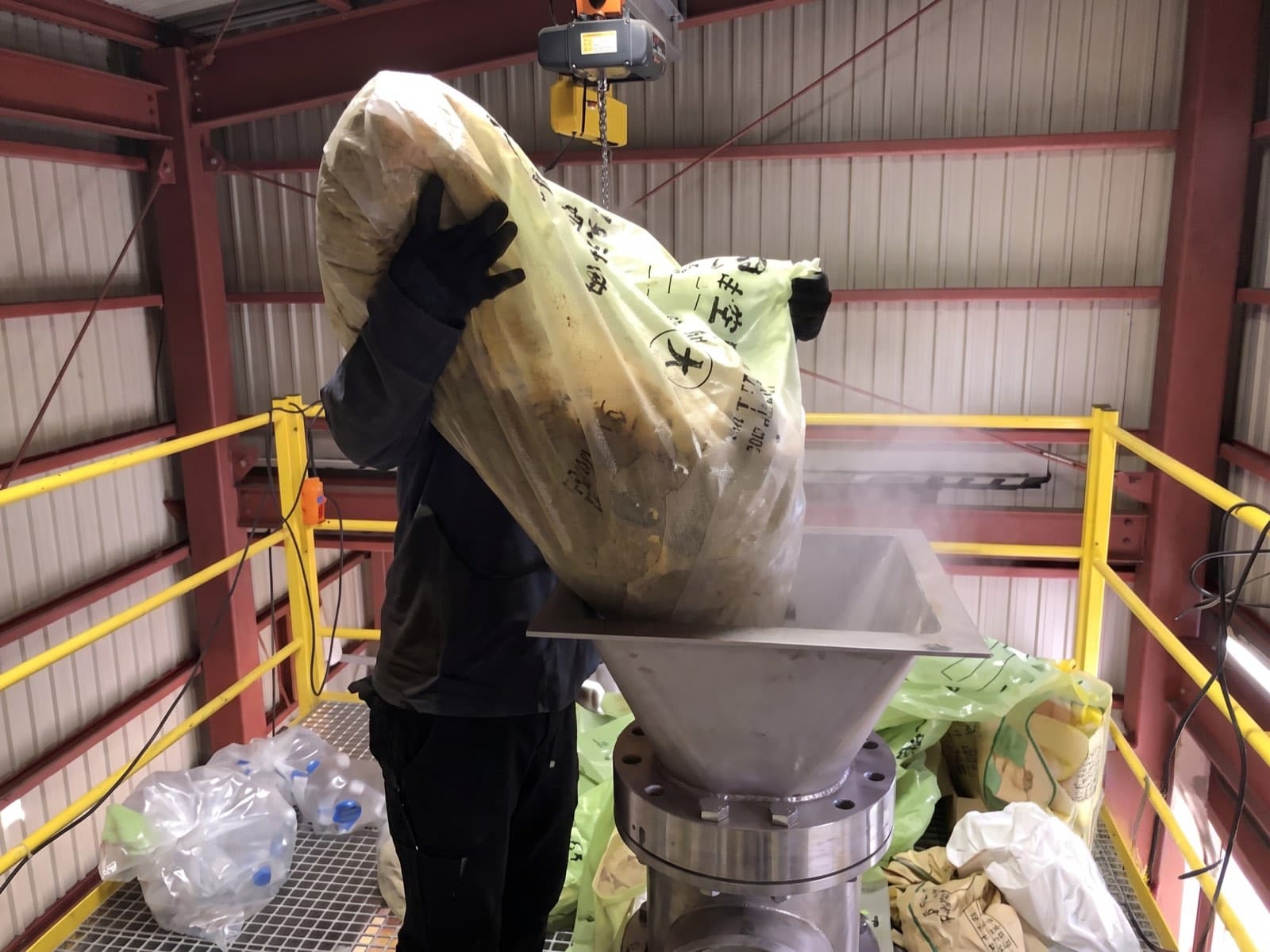 Operator lifting a mesh sack of waste into the stainless steel intake hopper of the PHANTOM system, steam rising from the sealed feed port — the single-step route that replaces three-stage cellulosic pretreatment before saccharification.