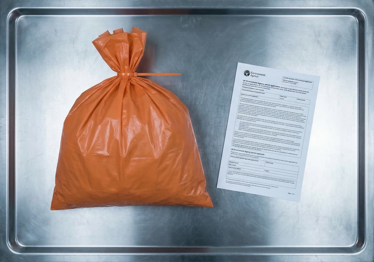 Orange clinical waste bag beside EA permit application document on stainless steel surface — the two physical objects UK healthcare operators must manage to achieve compliant on-site medical waste treatment under HTM 07-01
