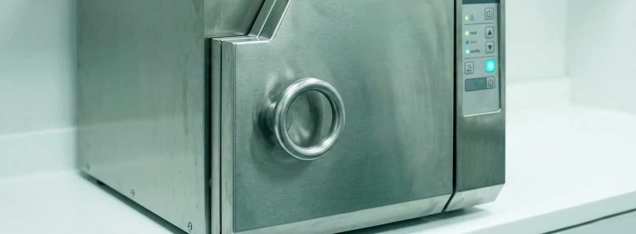 Bench-top hospital autoclave steriliser with closed porthole door in a clinical sterile services room — sterilises waste but cannot decompose it, reduce its classification, or eliminate downstream disposal costs